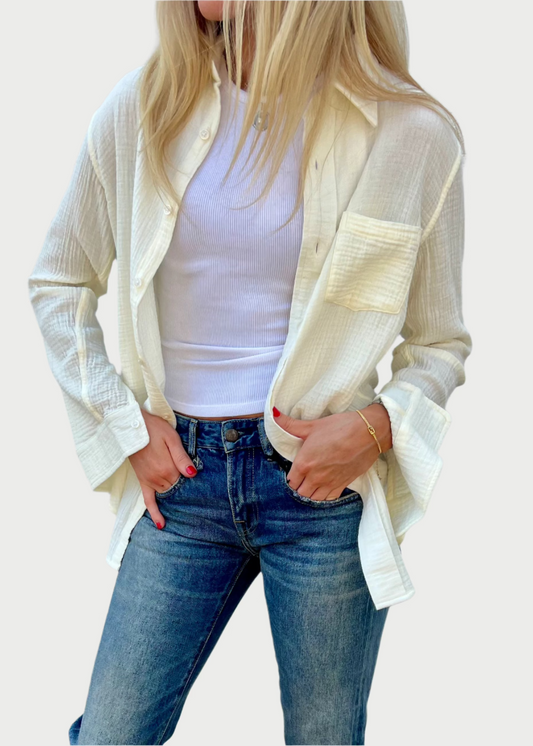 A person with long blonde hair stands against a white wall, wearing a white tank top, an oversized Denimist Boyfriend Crinkle Shirt, and blue jeans with hands casually placed in their pockets. They are accessorized with a thin Dylan James Jewelry bracelet and sport red nail polish.