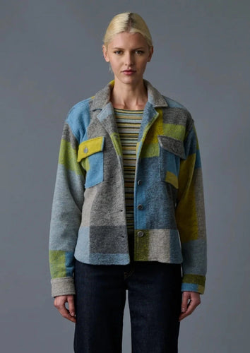 A person with blonde hair is wearing the Nirvana Pendleton Jacket by Le Superbe, a multicolored plaid design featuring shades of blue, gray, green, and yellow, paired with a striped shirt. This fall staple stands out beautifully against the plain gray background as they look directly at the camera.