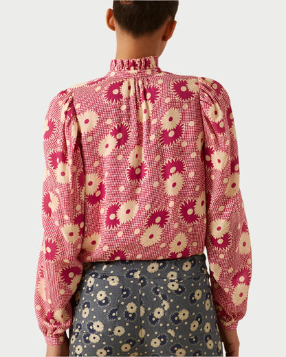 A person with short hair is depicted from behind, wearing the Annabel Berry Daisy Shirt by Alix of Bohemia, a vintage-inspired piece adorned with a red and white floral pattern and a high collar. They are also donning patterned pants with a darker design featuring a similar floral motif.