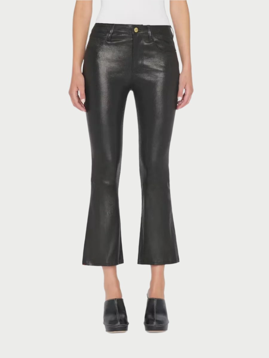 A person is standing against a plain white background, dressed in Frame's Le Crop Mini Boot Leather Pants and black shoes. The versatile pants, featuring a button and zipper closure, are made of shiny black leather. The person poses with their hands by their sides.