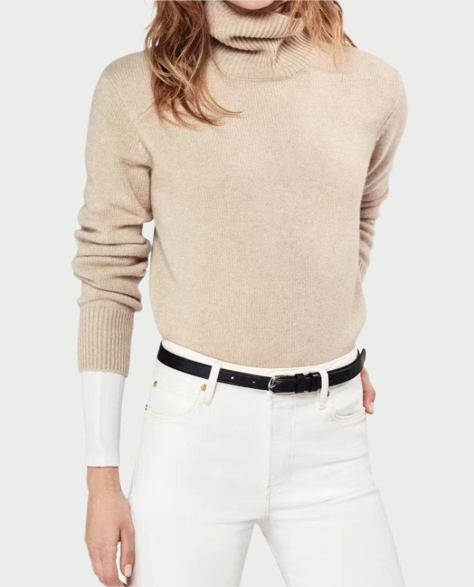 A person wearing an Oatmeal Cashmere Turtleneck Sweater by Frame paired with white high-waisted jeans and a black belt. The image focuses on the upper body, with arms relaxed by the sides, embodying a classic fall wardrobe against a plain off-white background.