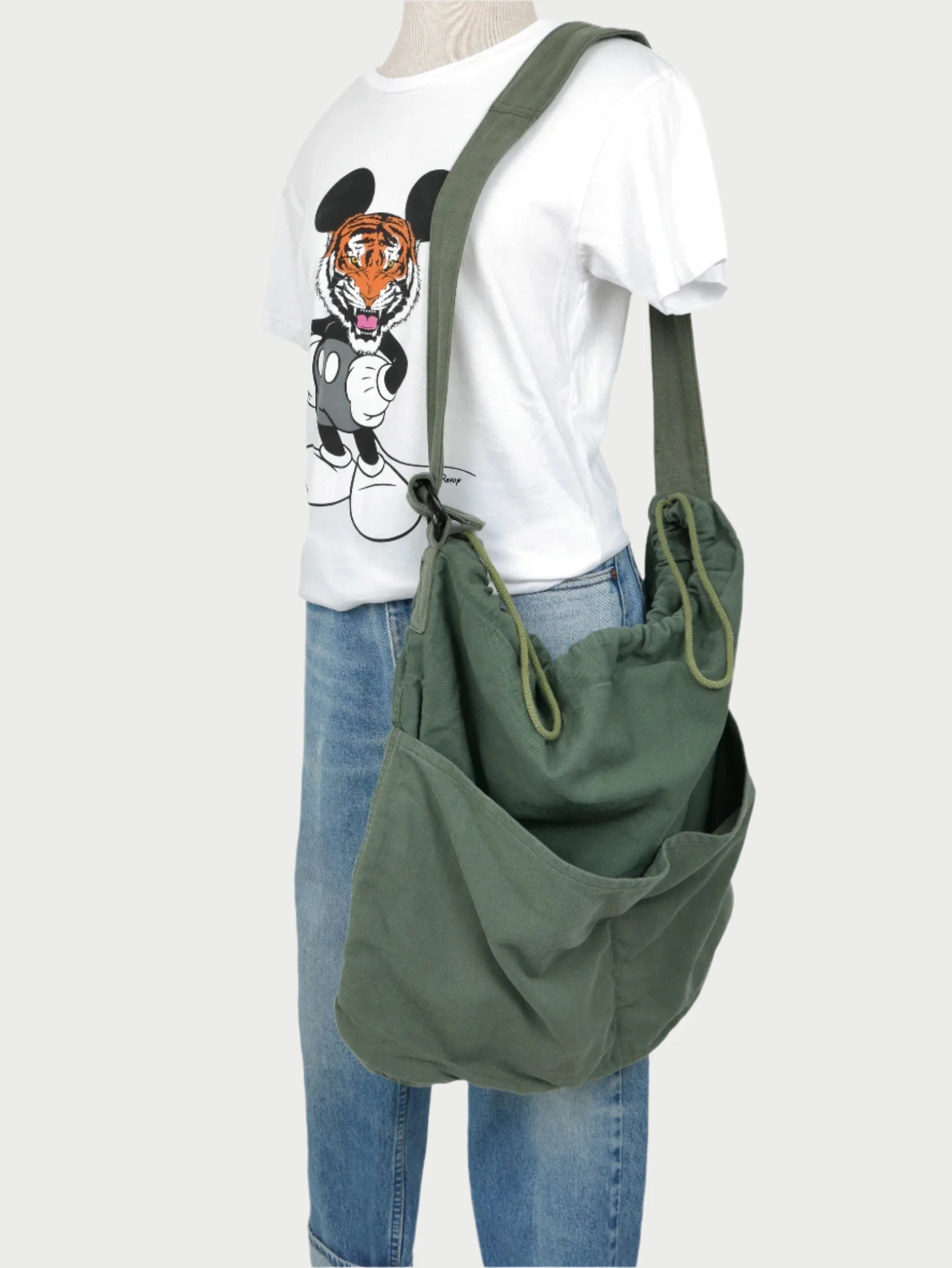 A mannequin showcases a white T-shirt featuring a tiger and ears design, paired with blue jeans. It is accessorized with the SUPER READY Drawstring Tote, an up-cycled Vintage US Military shoulder bag that includes an adjustable shoulder strap for versatility, casually slung over its left shoulder.