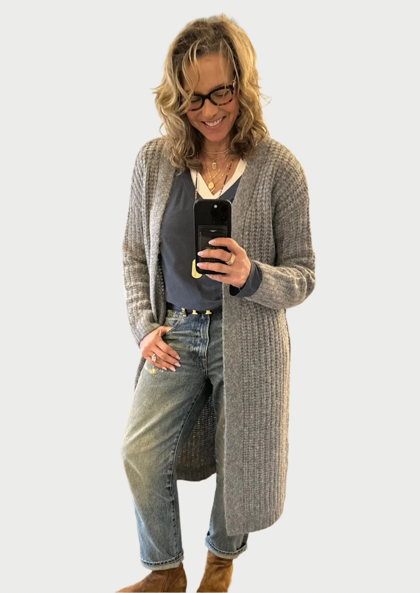 A woman with wavy hair and glasses takes a selfie in a clothing store. She is wearing a One Grey Day Chandler Duster, black top, jeans, and brown boots. The store has a stylish, cozy sophistication with pendant lights and clothes neatly displayed in the background.