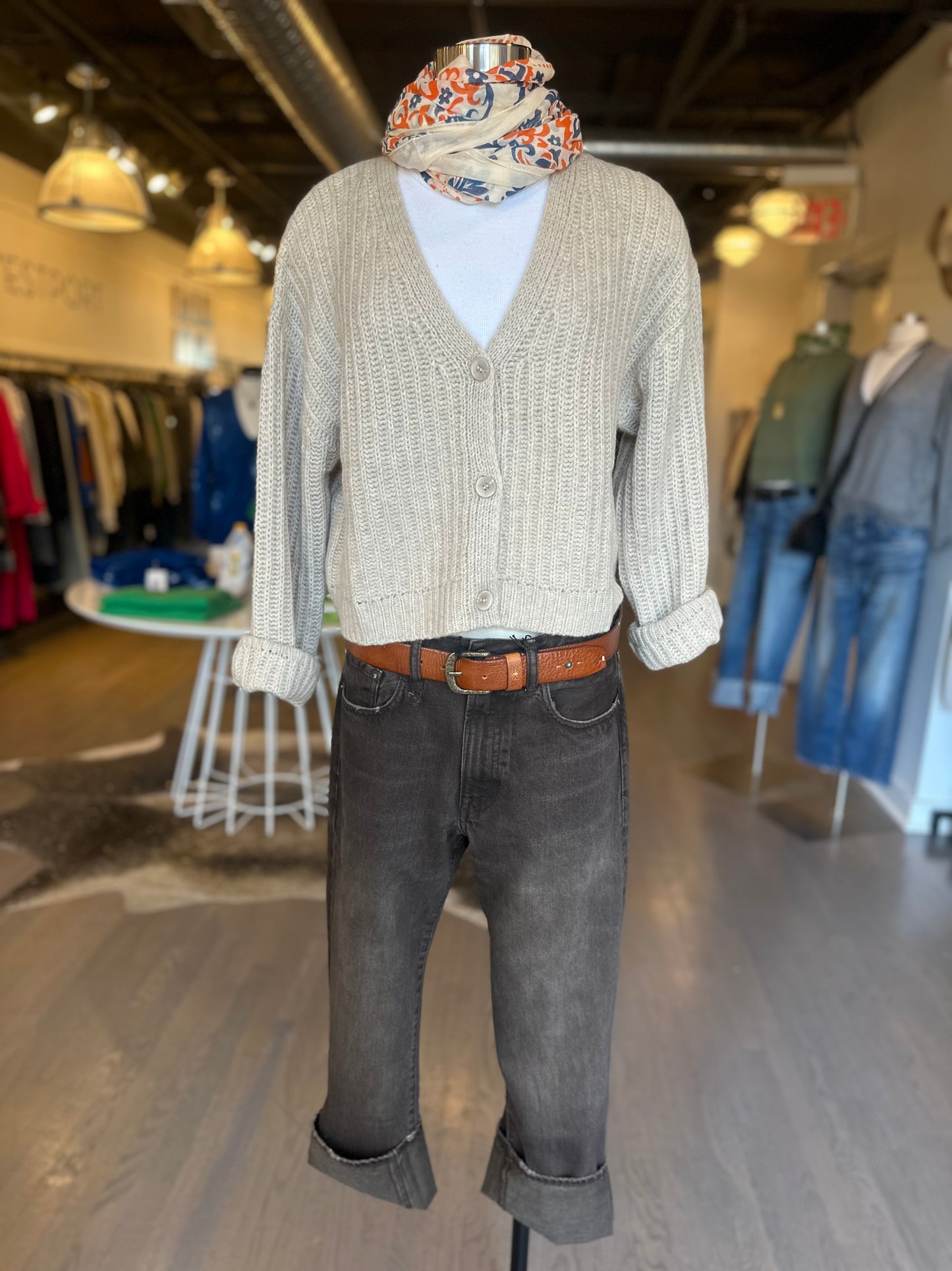 A mannequin displays the Polly Cashmere Cardigan in beige from One Grey Day over a white top, matched with rolled-up gray jeans and a brown belt. A vibrant scarf is elegantly wrapped around the neck, set against a backdrop of neatly arranged clothing on racks in the brightly lit One Grey Day store.