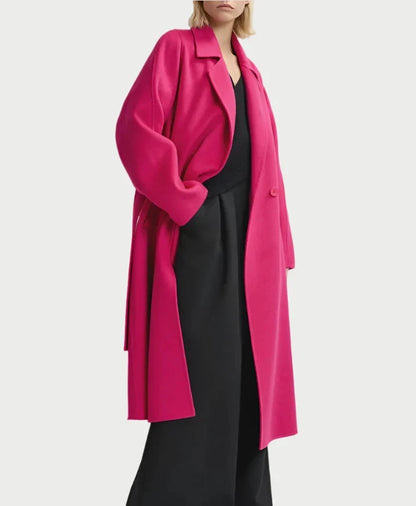 A person stands confidently against a plain white background, wearing the Essentiel Antwerp Double Face Coat in fuchsia with a matching belt and black wide-legged pants. The individual has short blonde hair and poses with one hand in their pocket.