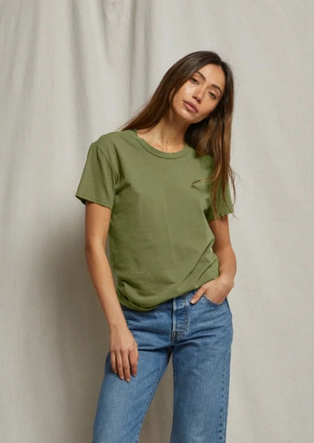 A person with long hair stands against a neutral fabric backdrop, wearing a green Harley - Safari T-shirt from perfectwhitetee and blue jeans. They pose with one hand in a pocket and look into the camera with a neutral expression, showcasing the timeless essential appeal of their outfit.