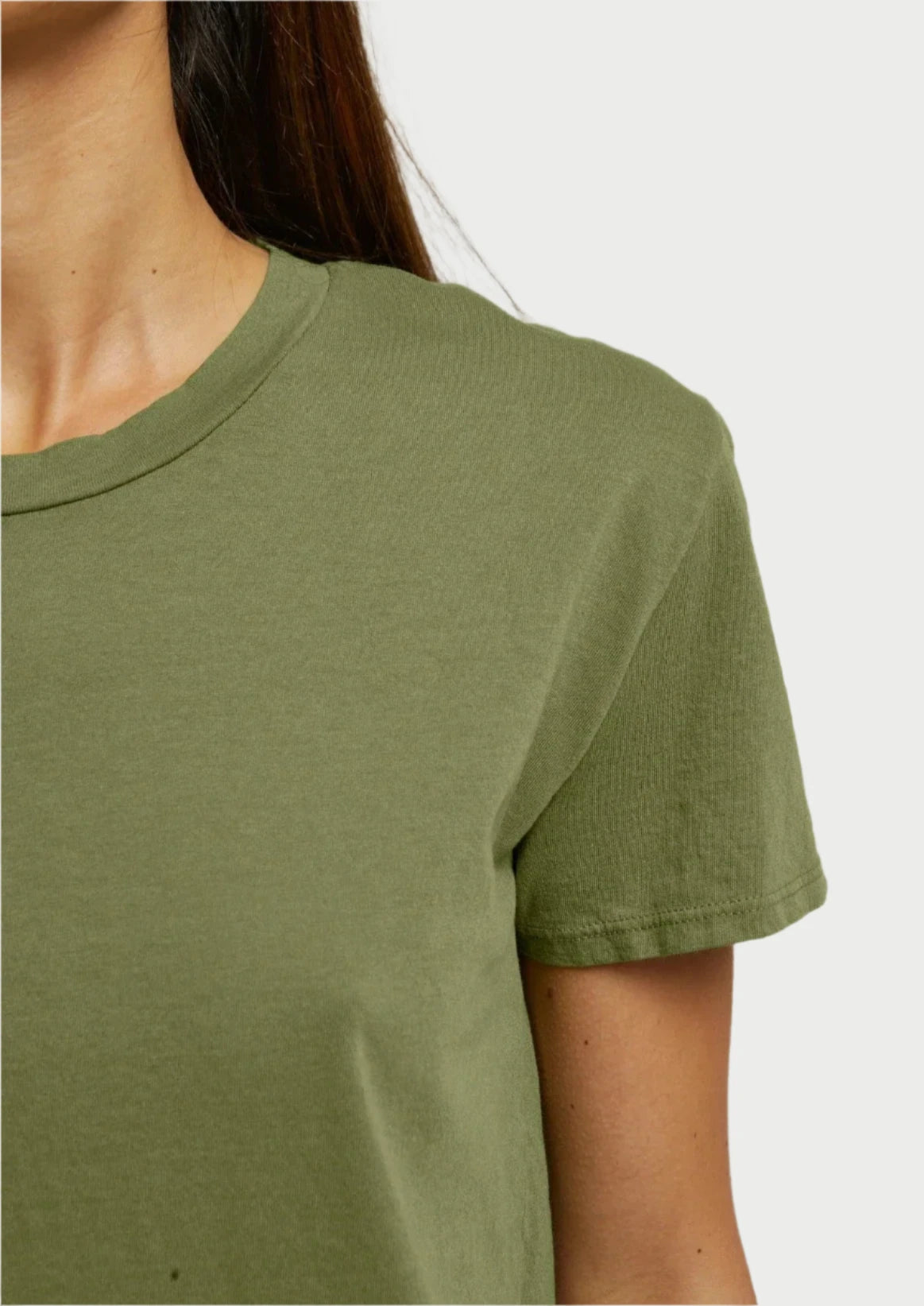 Close-up of a person wearing the Harley Tee Safari by perfectwhitetee, featuring a plain olive-green design with a comfortable fit. The focus is on the upper torso and right shoulder, highlighting its short sleeve and round neckline. The background is off-white, and the person's head is partially visible, with long brown hair cascading over their shoulder.