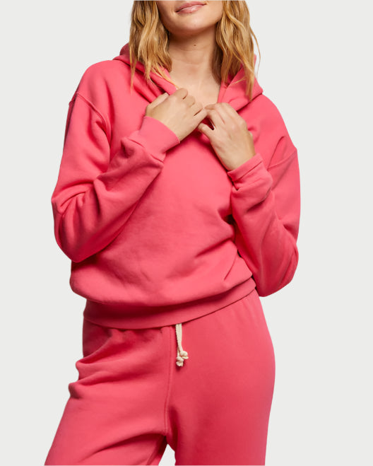 Wearing the Heart Hoodie from perfectwhitetee with matching pants against a plain background, a person with wavy hair holds the hoodie strings. The pink cotton fleece ensemble showcases a relaxed and casual fit, exuding effortless style and comfort.