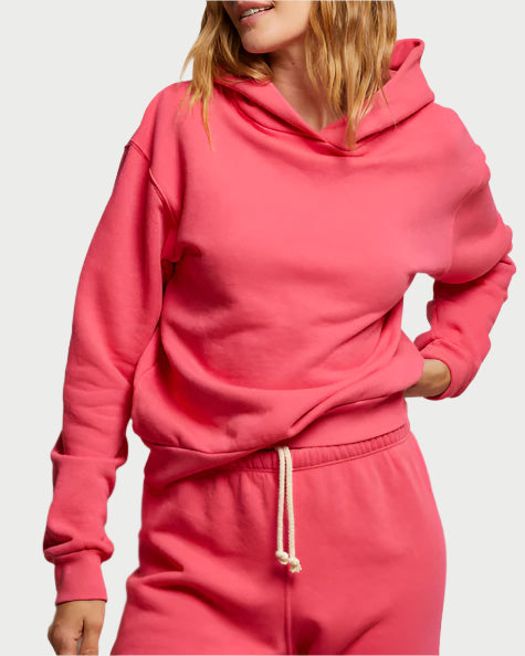 A person in a perfectwhitetee Heart Hoodie, bright pink cotton fleece with white drawstring, and matching sweatpants holds one hand in their pocket. The shot captures them from shoulders to waist against a light gray background.