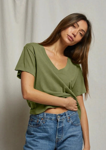 A woman with long brown hair, dressed in a flattering Hendrix- Safari tee by perfectwhitetee and blue jeans, stands against a plain backdrop. Slightly tilting her head, she holds the hem of her shirt with her right hand and wears a relaxed expression, epitomizing effortless everyday wear.