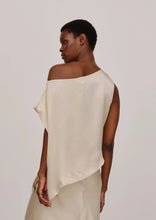 Load image into Gallery viewer, A person with short hair is seen from the back wearing a Herskind One Shoulder Silk Top in a cream-colored, off-shoulder asymmetrical design with a smooth texture. The background is plain and light grey.