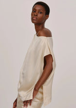 Load image into Gallery viewer, A person with short hair stands sideways facing the camera, wearing a loose, one-shoulder silk top. The One Shoulder Silk Top by Herskind has a satin-like texture and relaxed fit. The person looks ahead with a neutral expression against a plain, light-colored background.