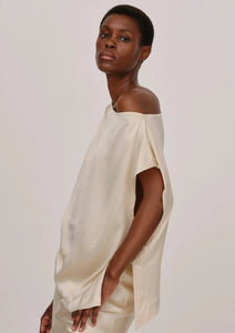 A person with short hair stands sideways facing the camera, wearing a loose, one-shoulder silk top. The One Shoulder Silk Top by Herskind has a satin-like texture and relaxed fit. The person looks ahead with a neutral expression against a plain, light-colored background.