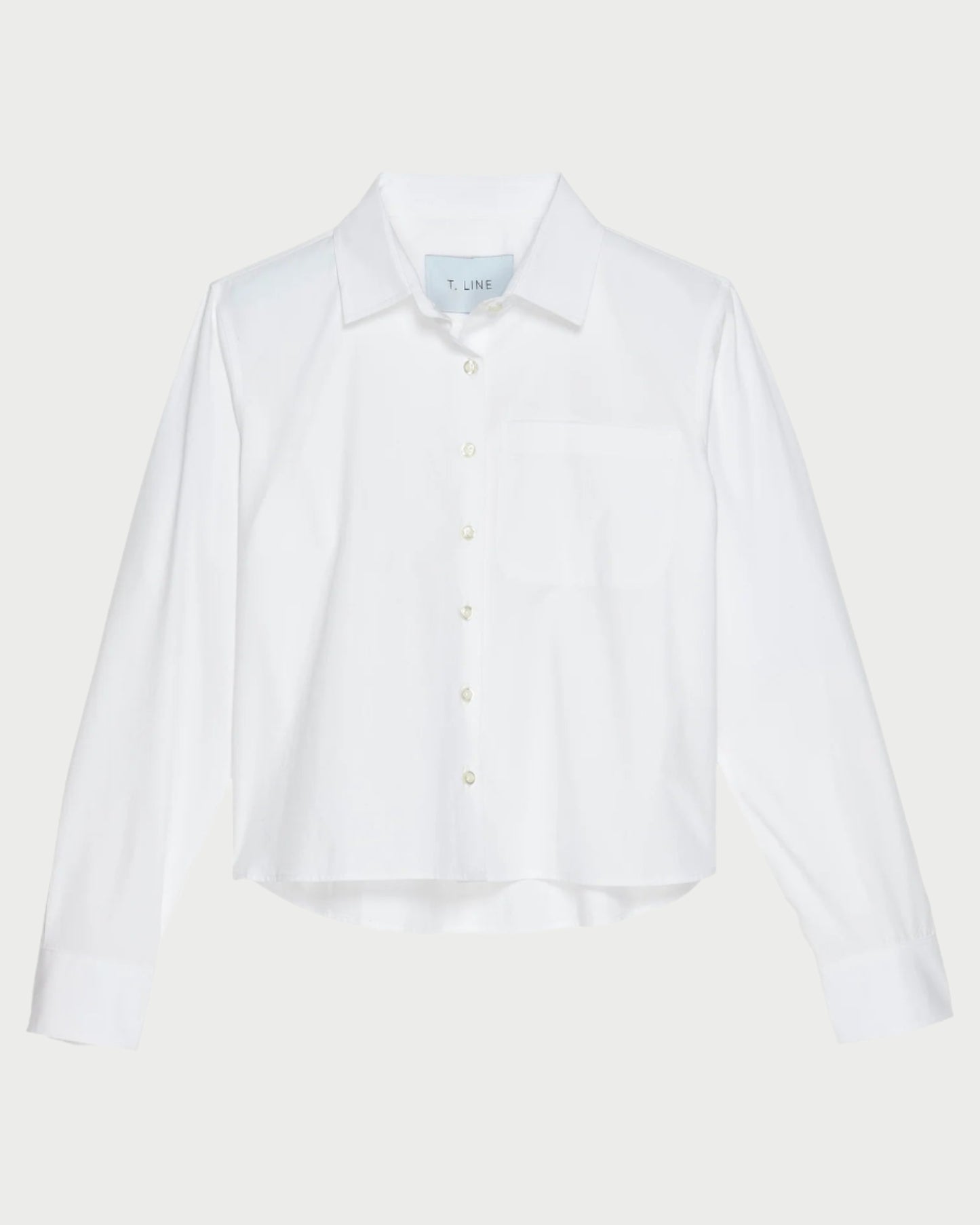 The T. Line Coco Cropped Shirt is a chic white long-sleeved button-up, featuring a stylish chest pocket and pointed collar. Made from 100% cotton, it stands gracefully against a light gray background.