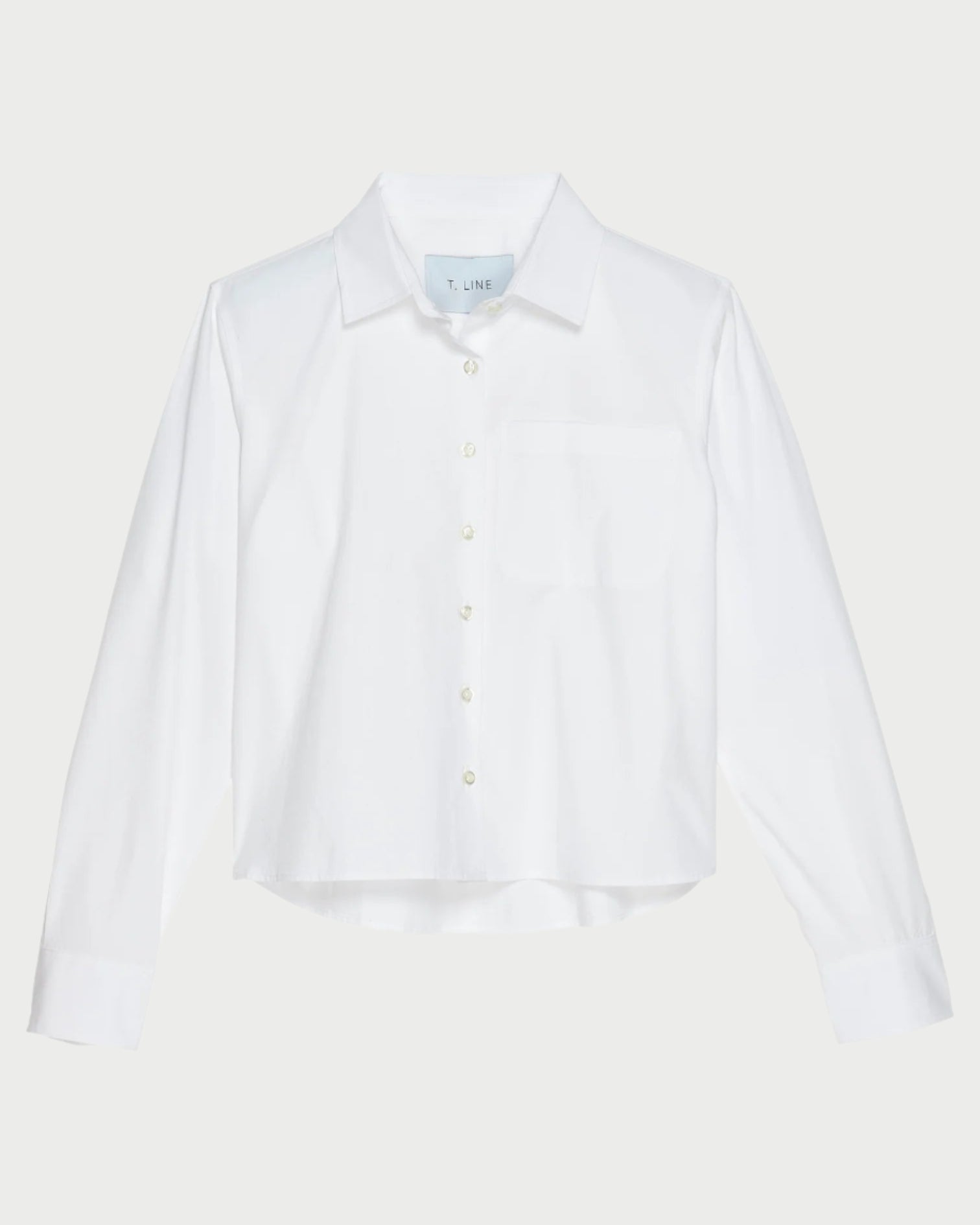 The T. Line Coco Cropped Shirt is a chic white long-sleeved button-up, featuring a stylish chest pocket and pointed collar. Made from 100% cotton, it stands gracefully against a light gray background.