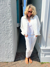 Load image into Gallery viewer, A person with curly blonde hair, wearing sunglasses, a white denim jacket over a Slim Denim Shirt by RE/DONE, white pants, and orange sandals, stands on a brick path between white and teal walls. The person is smiling with hands in their pockets, enjoying a sunny day.