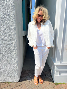 A person with curly blonde hair, wearing sunglasses, a white denim jacket over a Slim Denim Shirt by RE/DONE, white pants, and orange sandals, stands on a brick path between white and teal walls. The person is smiling with hands in their pockets, enjoying a sunny day.