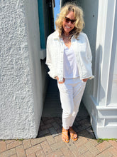 Load image into Gallery viewer, A smiling woman with curly blonde hair and sunglasses stands in a narrow walkway between white buildings. She is wearing the Slim Denim Shirt from RE/DONE under a white denim jacket, white pants, and brown sandals. Light from above casts gentle shadows on the textured wall behind her.