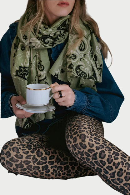 A person with long hair, wearing a Maison Lecomte Flament Large Cotton Voile Scarf, denim jacket, and leopard-print leggings, sits on a bed holding a cup of coffee on a saucer. A Krispy Kreme box is on the bed near them. The setting appears relaxed with a neutral background.