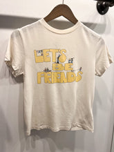 Load image into Gallery viewer, The Lets Be Friends Classic Tee from RE/DONE is a versatile white short-sleeved T-shirt hanging on a wooden hanger. The front showcases &quot;LET&#39;S BE FRIENDS&quot; in large, yellow letters, adorned with small illustrations of cartoon children in various poses interacting with the text, making it a playful addition to any wardrobe.