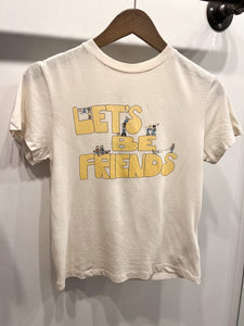 The Lets Be Friends Classic Tee from RE/DONE is a versatile white short-sleeved T-shirt hanging on a wooden hanger. The front showcases "LET'S BE FRIENDS" in large, yellow letters, adorned with small illustrations of cartoon children in various poses interacting with the text, making it a playful addition to any wardrobe.