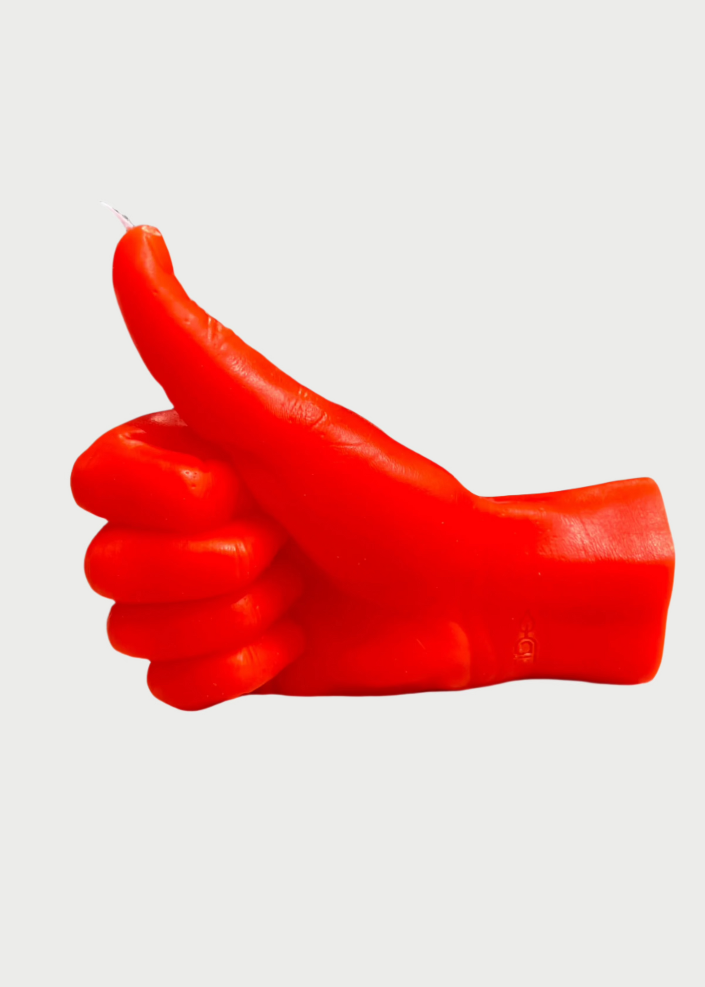 The 54° Celsius Thumbs Up Candle, a vibrant red hand-shaped candle on a light gray surface, vividly displays its color and character. Molded to depict a thumbs-up gesture, it stands out against the slightly blurred outdoor backdrop.