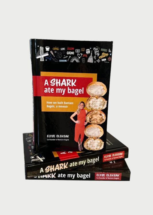 A stack of books titled "A Shark Ate My Bagel" by Elyse Oleksak are displayed on a shelf. The cover shows a photo of a woman in a red dress next to three Bantam Bagels. In the background, a purse and other items are visible, capturing the essence of this inspirational memoir featured on Shark Tank.