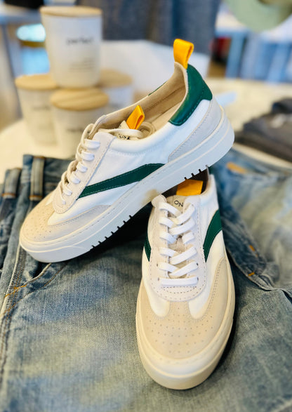 A pair of stylish Oncept Lagos Shoes with vibrant green accents and yellow pull tabs are displayed on top of a pair of blue jeans. In the background, several coffee cups and blurred objects enhance the contemporary style of the scene.