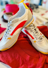 Load image into Gallery viewer, A pair of Oncept Montreal Shoes with red and yellow accents are placed on top of a red t-shirt. The shoes, featuring a sleek design with grey laces and a white body, exude effortless style. The setting is a brightly lit room with various indistinct items blurred in the background.