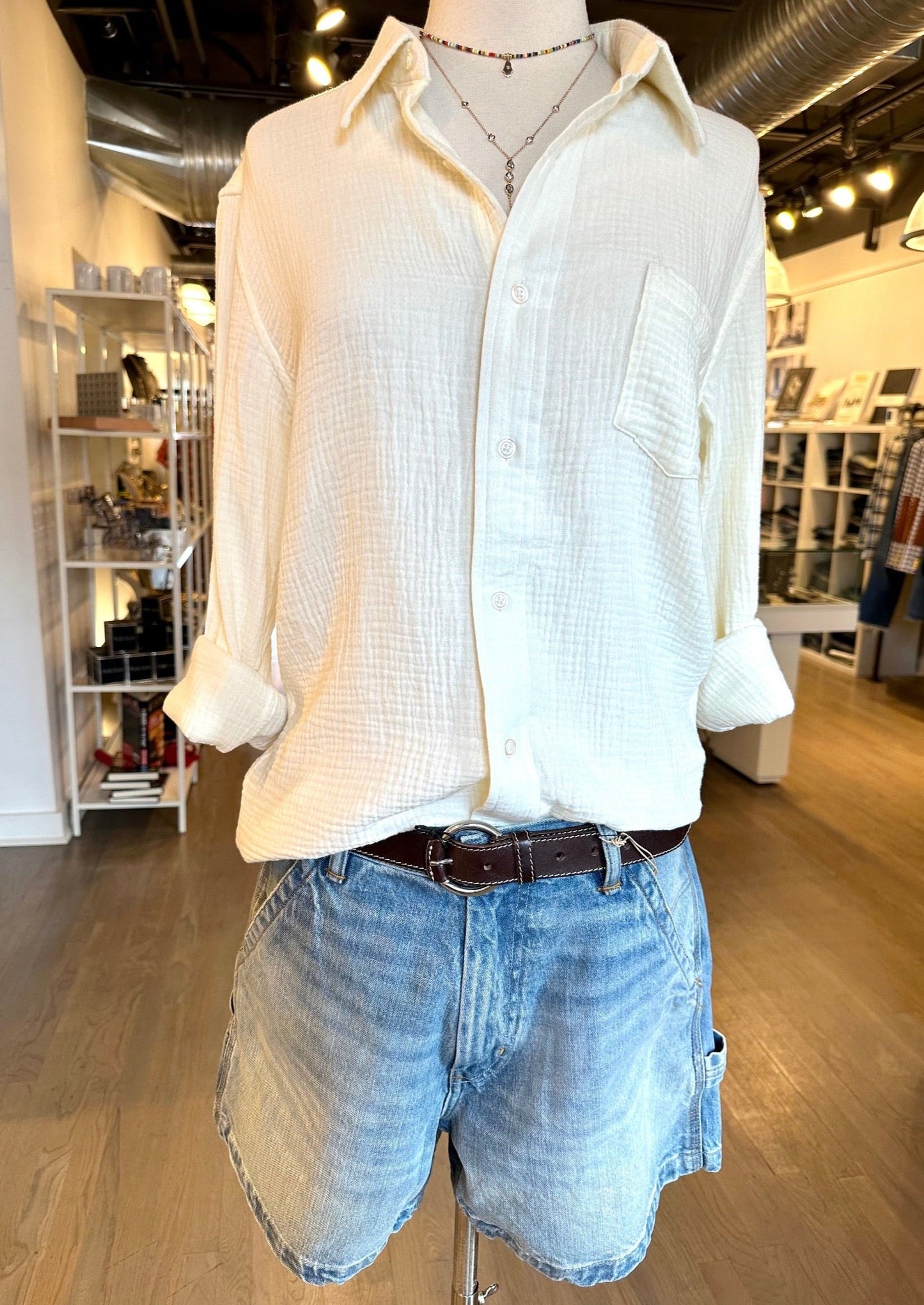 A mannequin in a clothing store is wearing a light, textured white Denimist Boyfriend Crinkle Shirt with rolled-up sleeves, tucked into a pair of casual light-wash denim shorts belted at the waist. The store background includes shelves and hanging garments from Dylan James Jewelry.