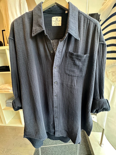 A dark blue, Button Front Shirt by Denimist with a single chest pocket is displayed on a wooden hanger. This summer piece exudes a relaxed, casual style with its textured fabric and rolled-up sleeves. Its lightweight design makes it perfect for warm weather. Shelves with folded clothes are visible in the background.