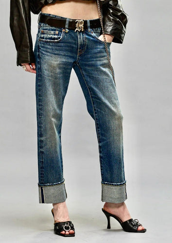 A person wears rolled-up, slim fit R13 Cuffed Boy Straight blue jeans paired with black heeled sandals. They have a black leather jacket and a belt with a prominent metallic buckle. The background is plain grey. Only the lower half of their body is visible.