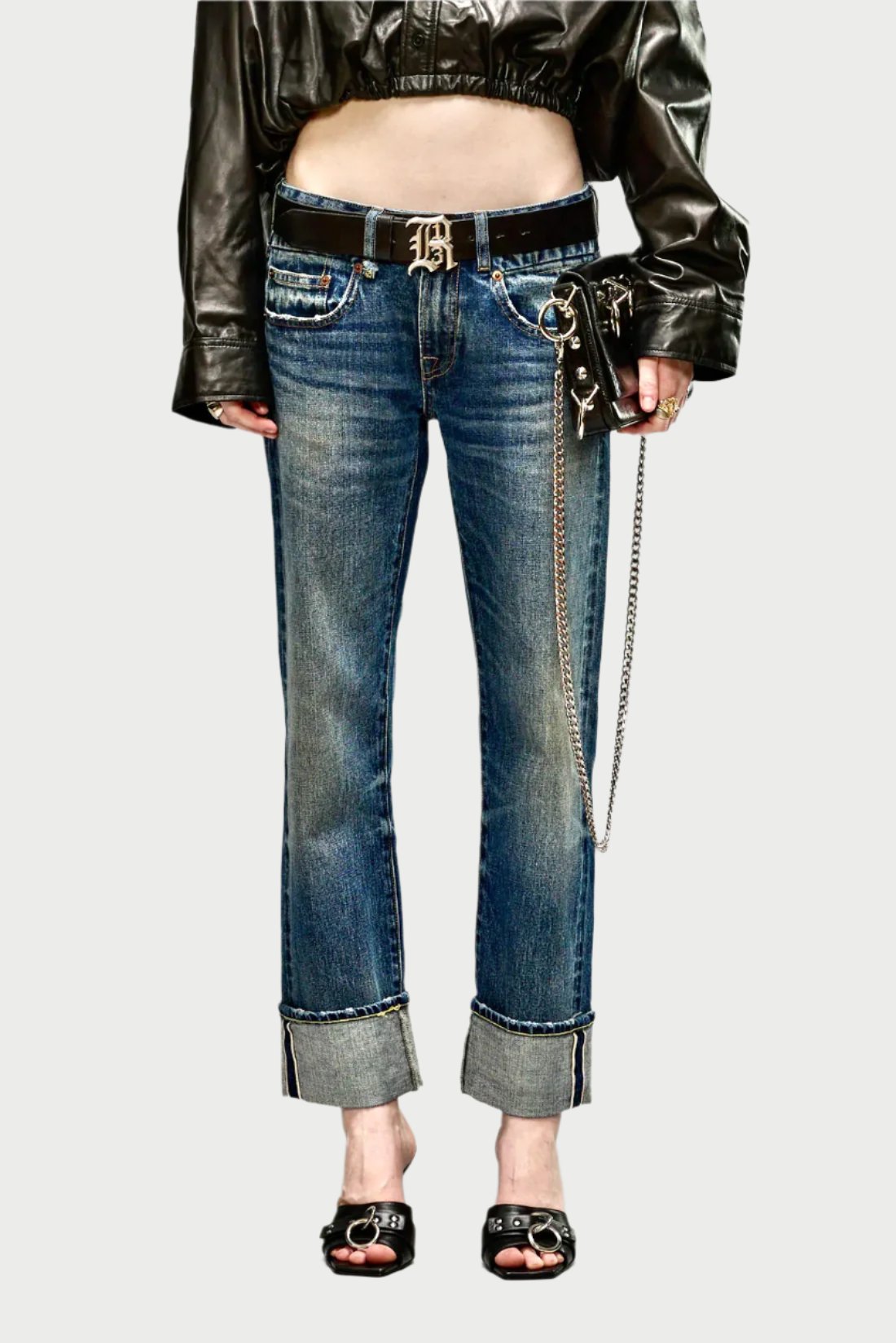 A person models cuffed, distressed R13 Cuffed Boy Straight jeans with a black leather belt showcasing a prominent "B" buckle. They wear a black leather cropped jacket and have a keychain and wallet attached to the belt. The outfit is completed with black open-toe sandals.