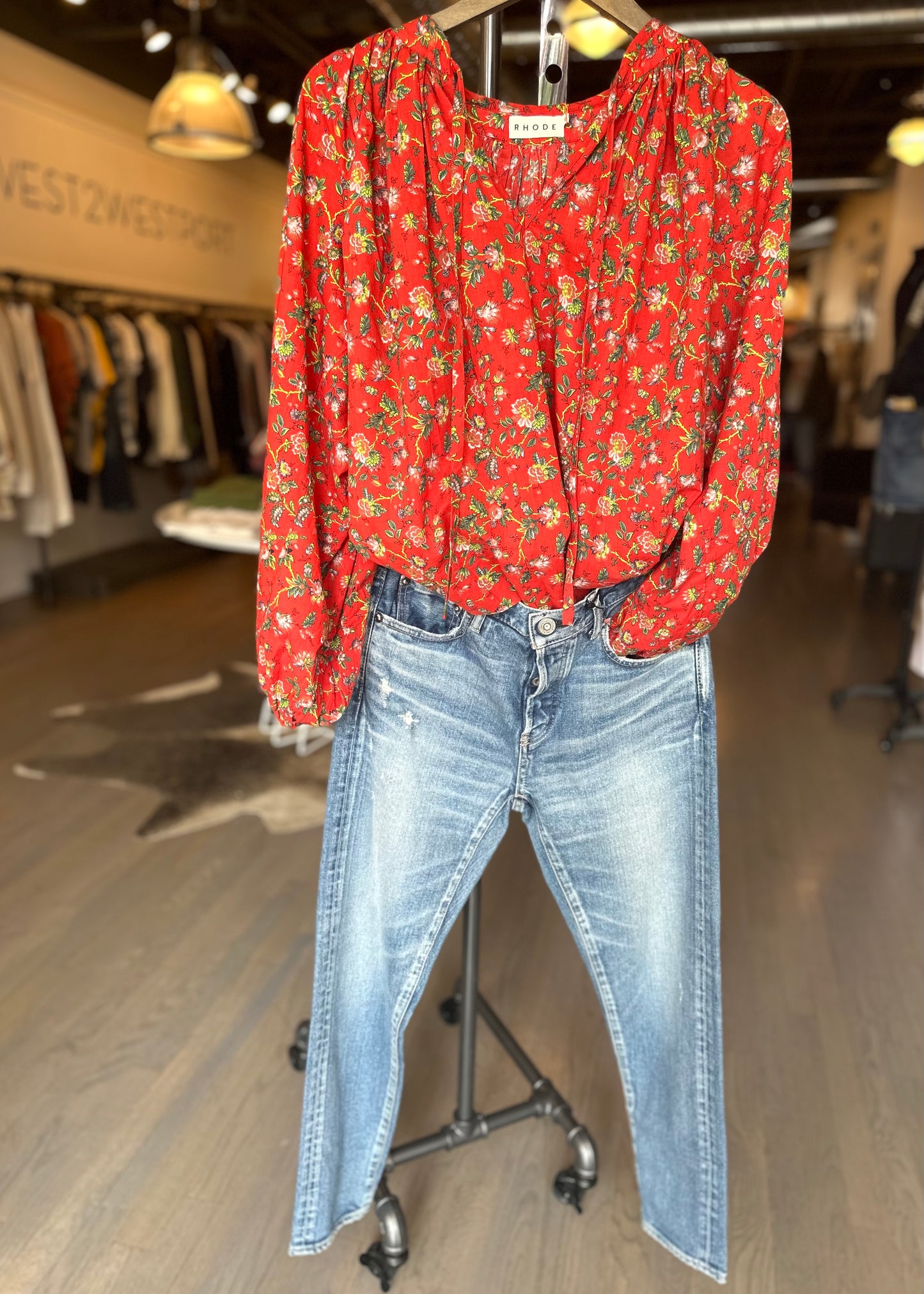 A mannequin is dressed in a Rhode Red Flowered Poet Top featuring a gathered neckline, paired with blue denim jeans. The outfit is displayed in a clothing store, surrounded by racks of clothing in the background. The store boasts wooden flooring and warm lighting.