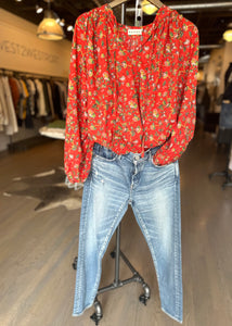 A mannequin is dressed in a Rhode Red Flowered Poet Top featuring a gathered neckline, paired with blue denim jeans. The outfit is displayed in a clothing store, surrounded by racks of clothing in the background. The store boasts wooden flooring and warm lighting.