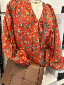 A mannequin is dressed in a Rhode Red Flowered Poet Top, featuring a V-neckline and long puffed sleeves adorned with a delicate flower pattern. The mannequin accessorizes with layered necklaces and carries a tan crossbody handbag.