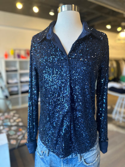 A mannequin showcasing a chic ink blue Tawny Sequin Top and light blue jeans stands prominently in a well-lit Zadig & Voltaire store, with shelves and clothing racks subtly enhancing the stylish backdrop.