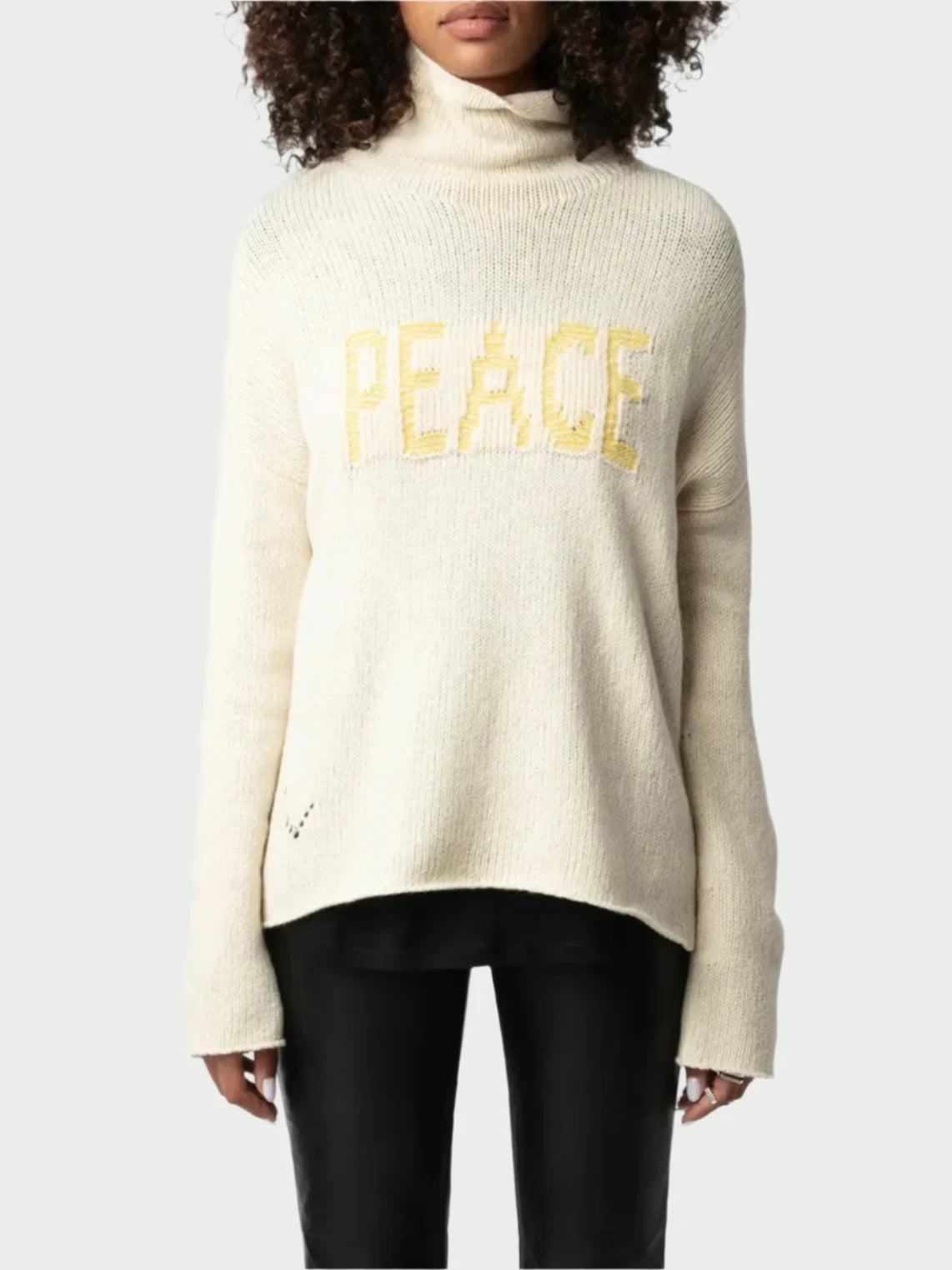 A person with curly hair wearing sunglasses is dressed in the Zadig & Voltaire Peace Sweater, a cream merino wool garment featuring the word "PEACE" in bold yellow letters on the front. They are also wearing black pants against a plain, light-colored background.