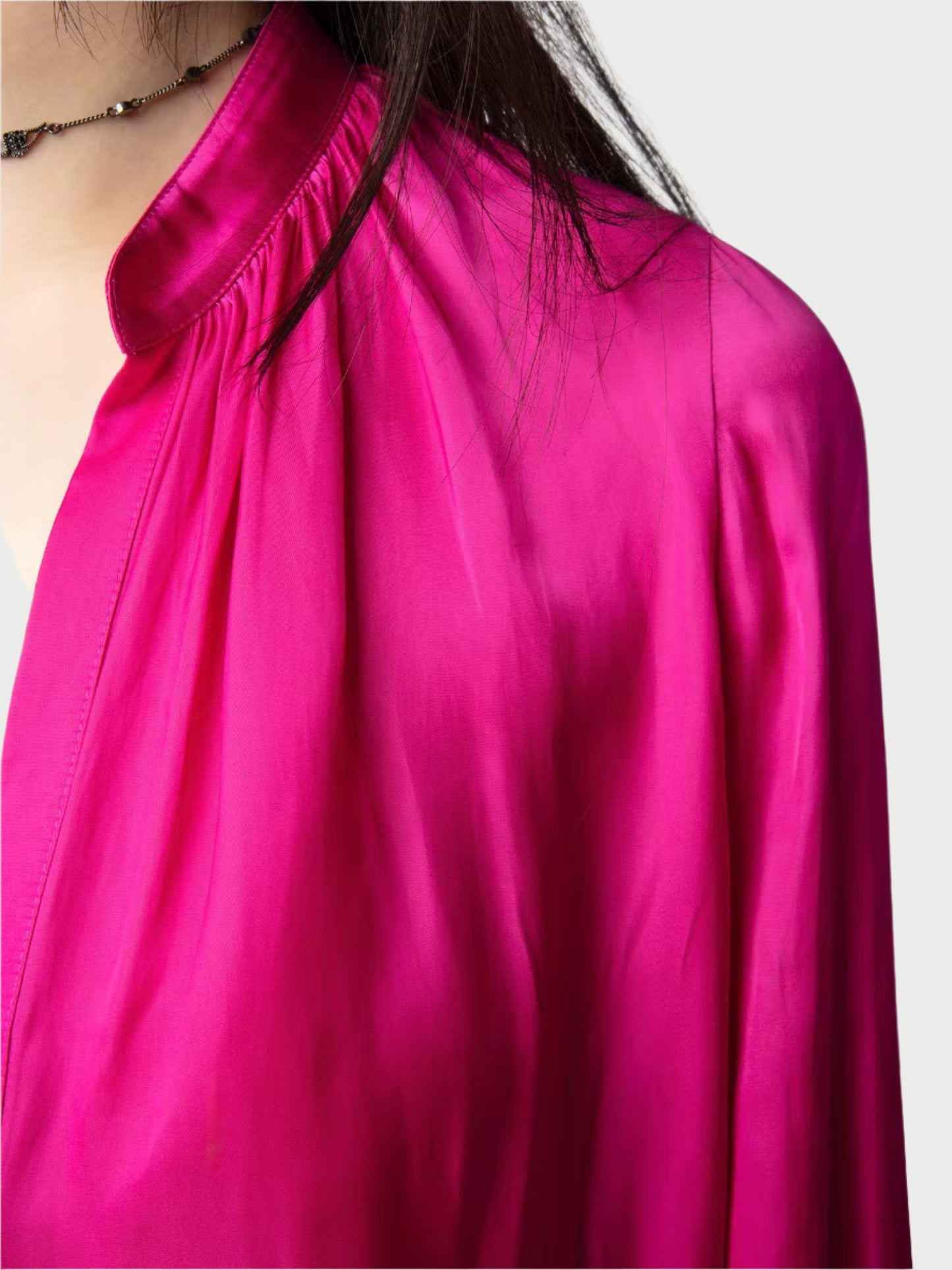 A person wearing a vibrant Bright Pink Satin Blouse from Zadig & Voltaire, featuring a high collar and gathered detailing on the shoulder. The blouse shimmers in the light, and the person's long dark hair partially covers their face. They are accessorized with a delicate black necklace.