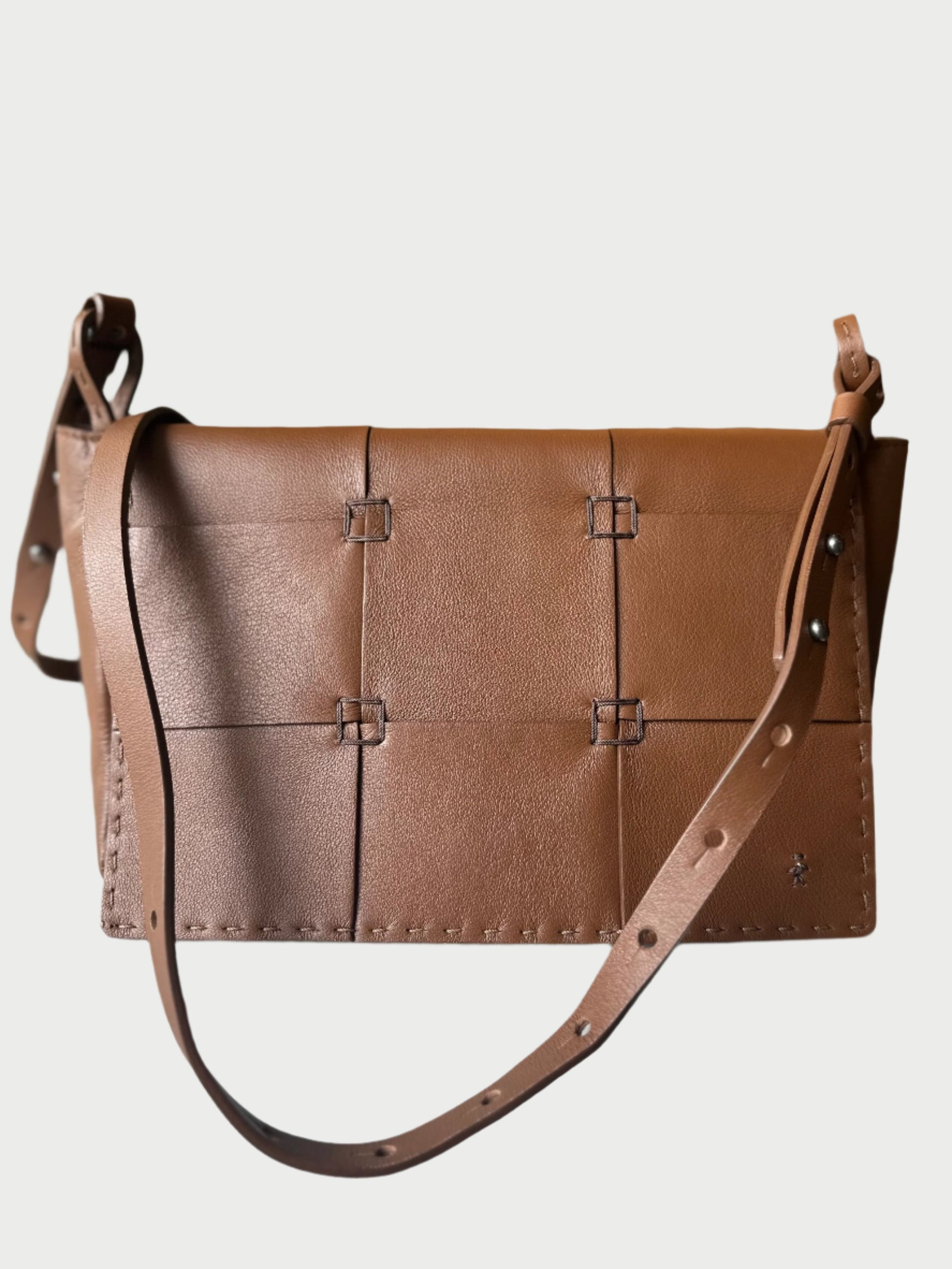 A Handmade Leather Patchwork Bag by Henry Beguelin is showcased on a white surface, featuring brown calfskin leather with an eye-catching handmade patchwork design. The bag includes a long adjustable strap and intricate decorative stitching. In the softly blurred backdrop, a peace sign composed of lights adorns the wall.