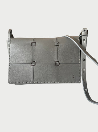 Displayed on a white surface is the Handmade Leather Patchwork Bag by Henry Beguelin, a stylish gray calfskin leather crossbody featuring stitched detailing and an adjustable strap with a flap closure. In the background, blurred shelves and decor elements can be seen.