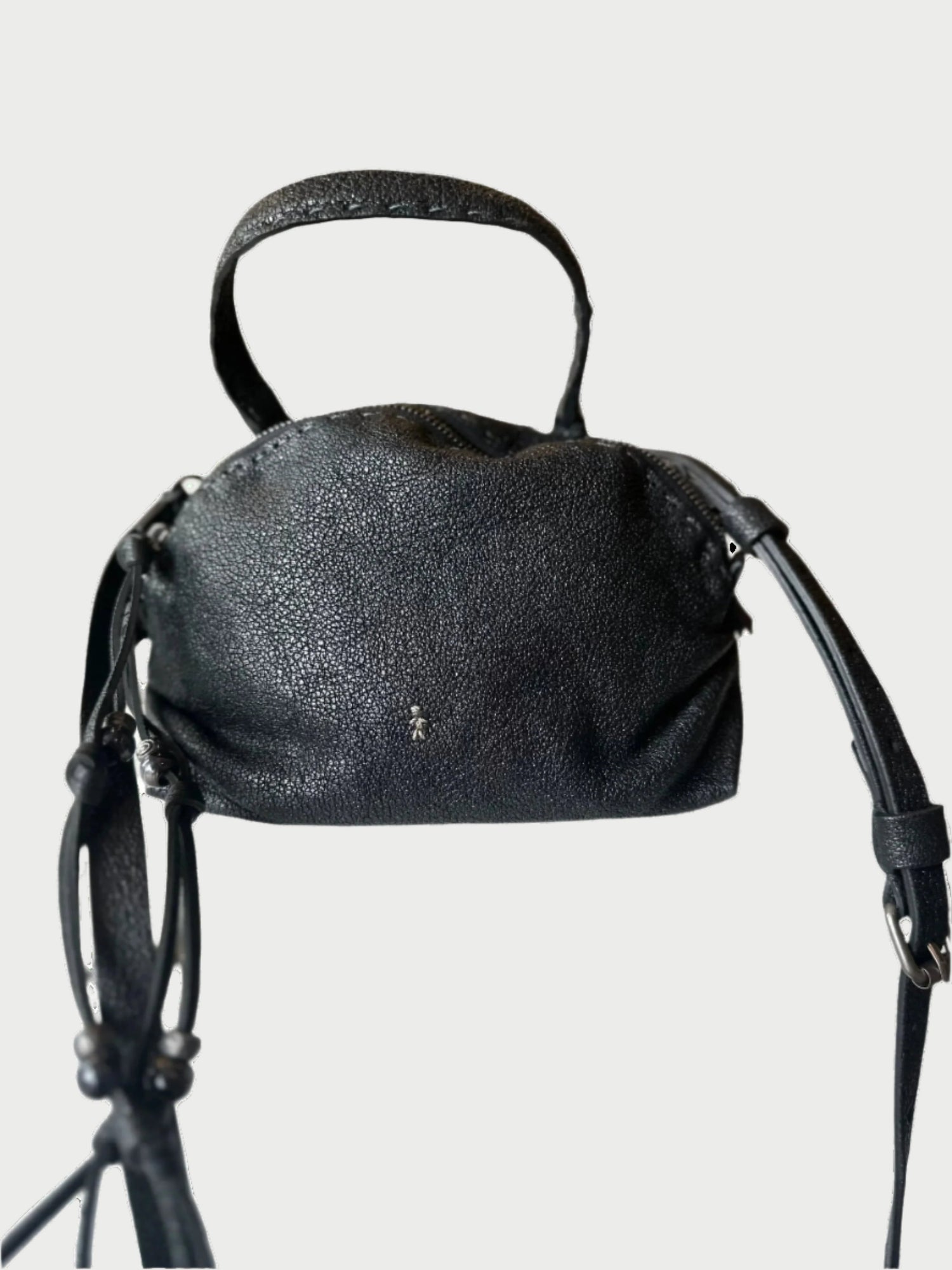 A black textured Henry Beguelin Vegetal Lamè Bag, with both a short handle and a long shoulder strap, is placed on a white surface. The softly blurred background highlights shelves filled with colorful items. Crafted from vegetable-tanned leather, the bag displays a small, subtle logo on the front.