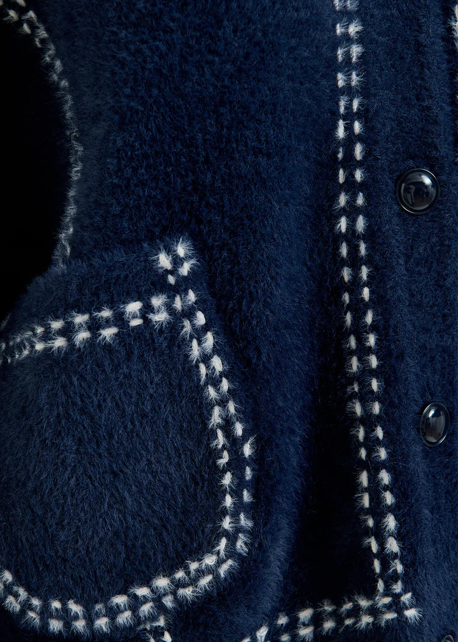 Close-up of Essentiel Antwerp's Contrasting Stitch Knit Cardigan, showcasing the textured blue knit with decorative white stitching, black buttons, a front pocket, and part of the button placket.