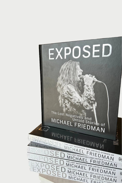 A stack of books titled "EXPOSED- The Lost Negatives" from Michael Friedman Photography is on display in a clothing store. Featuring never-before-seen images, the cover shows a black-and-white photo of a singer performing, with clothing racks and various garments visible in the background.