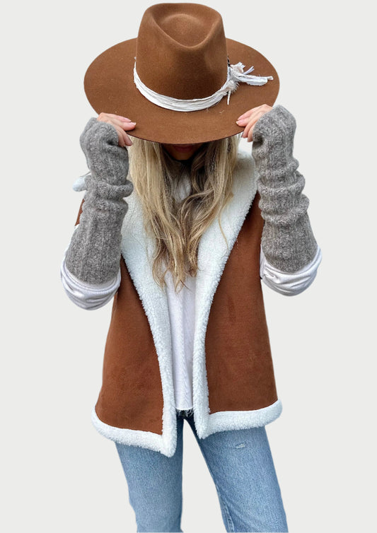 A person with long, blonde hair wears a brown hat with white rope detailing, a brown and white shearling vest, a white top, and luxurious Meg Cohen Hand Knit Arm Sox in gray. Their head is tilted down, partially covering their face with the hat brim. The background is a pale blue wall.