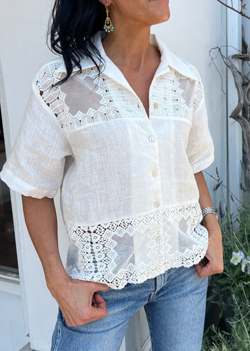 A person stands wearing a chic and breezy Alix of Bohemia Eyelet Shirt with intricate lace details on the shoulders and hemline. The person pairs the shirt with blue jeans and wears drop earrings. The background shows a white wall and some foliage, perfect for summer picnics.