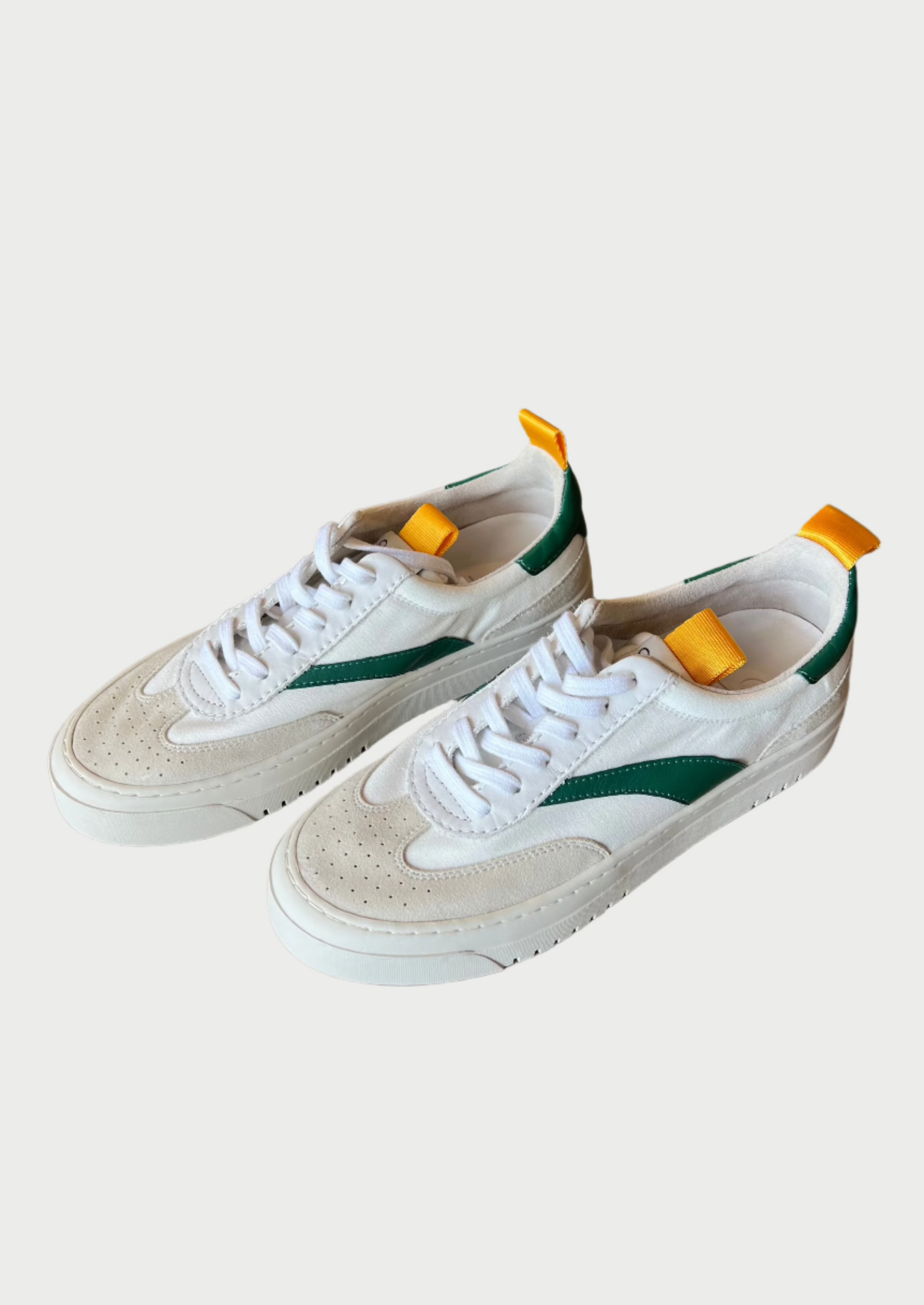 A pair of Lagos Shoes by Oncept in a contemporary style, featuring vibrant green detailing on the sides and orange pull tabs on the back, are placed on a wooden floor. The white shoes have matching laces and perforated toe boxes.