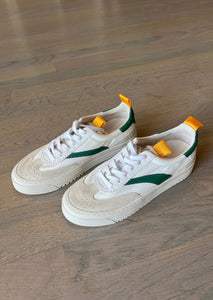 A pair of Lagos Shoes by Oncept in a contemporary style, featuring vibrant green detailing on the sides and orange pull tabs on the back, are placed on a wooden floor. The white shoes have matching laces and perforated toe boxes.