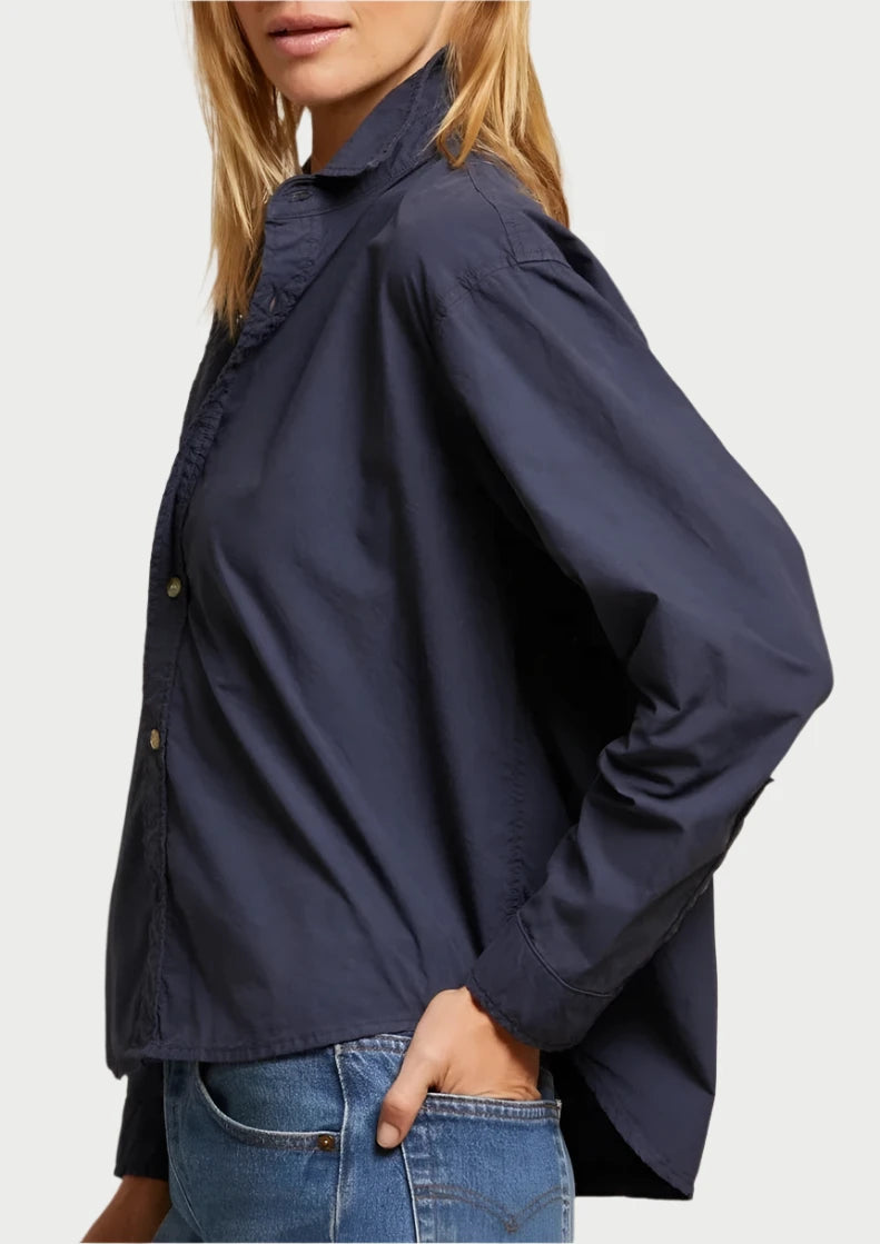 A woman with shoulder-length blonde hair stands sideways, looking at the camera. She is wearing a navy blue James Button Down Shirt from perfectwhitetee and blue jeans, exuding an effortlessly stylish vibe. Her neutral expression complements the plain, light-colored wall in the background.