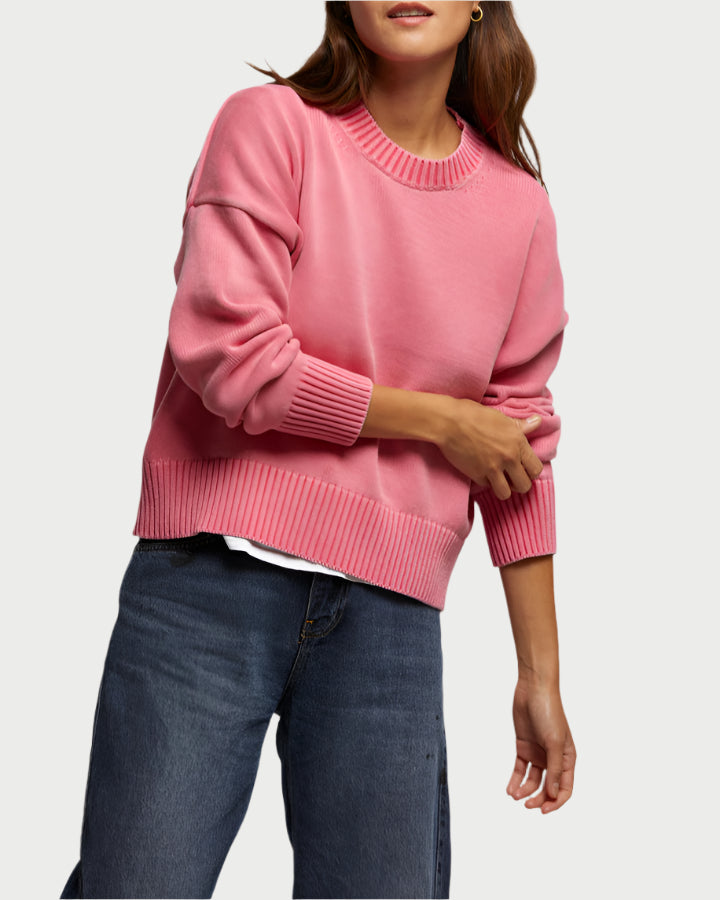 Donning the Janet Sweatshirt in pink by perfectwhitetee, a person wears it over a white shirt with blue jeans. With its boxy fit, their left arm is bent as they adjust the sleeve using their right hand against a plain white background.